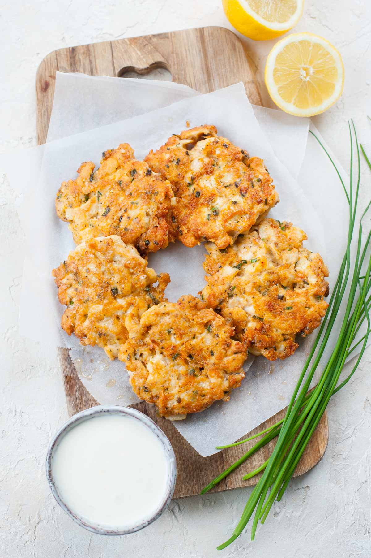 Cheesy chicken fritters on a wooden board, surrounded with chives and lemon halves.