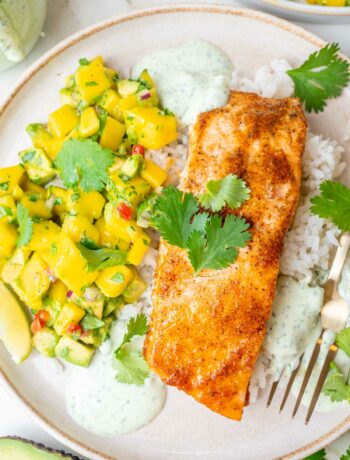 Baked salmon with mango avocado salsa, rice, and cilantro lime crema on a beige plate.