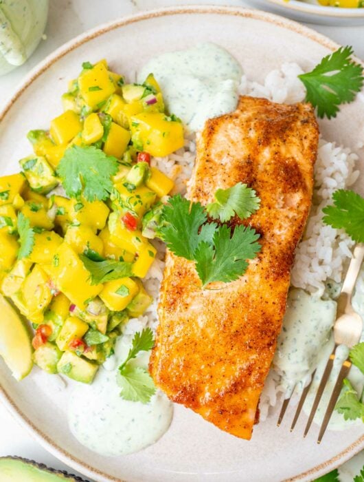 Baked salmon with mango avocado salsa, rice, and cilantro lime crema on a beige plate.