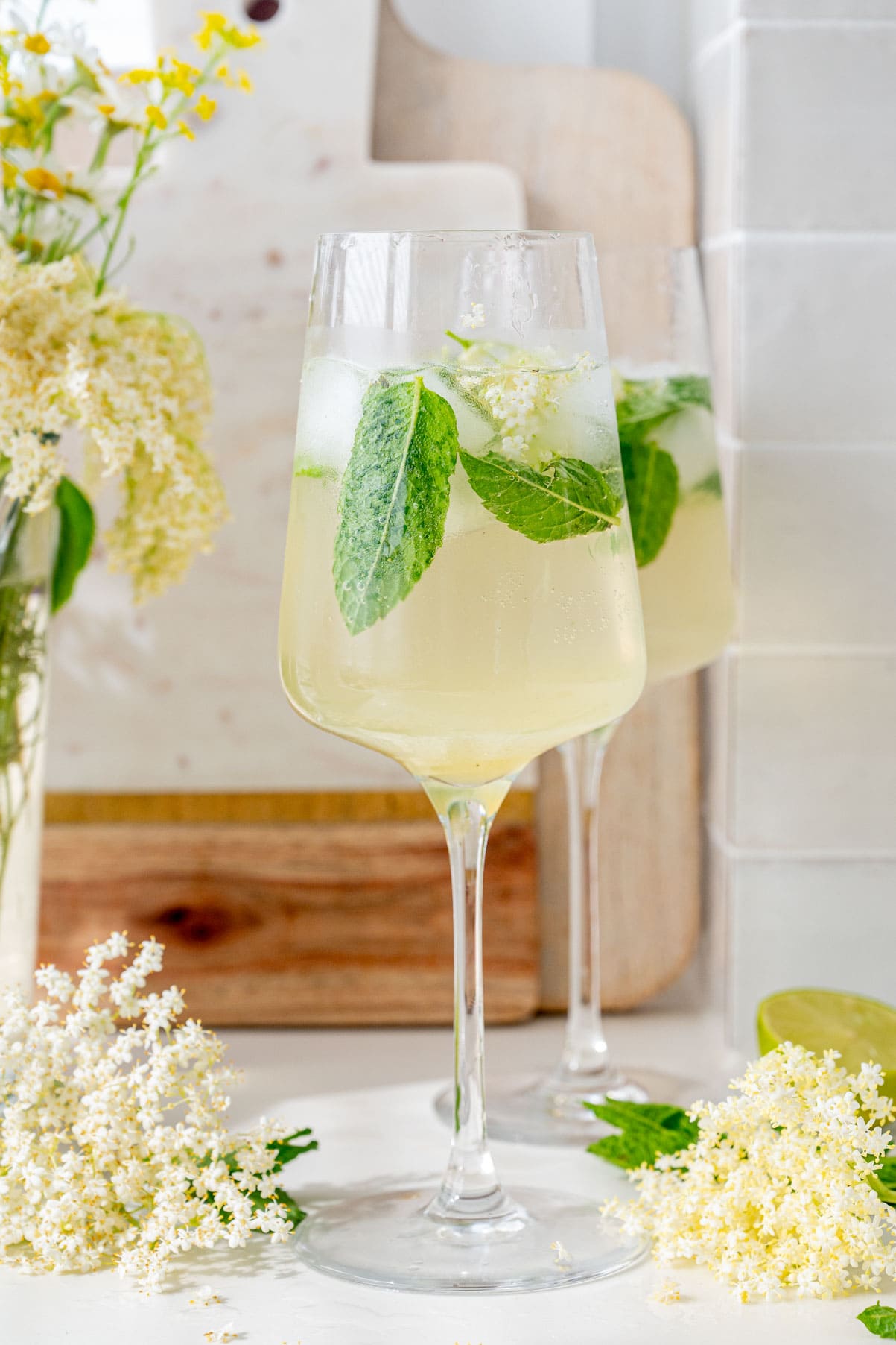 Two glasses with Hugo Cocktail. Elderflowers, mint leaves, and flowers in the background.