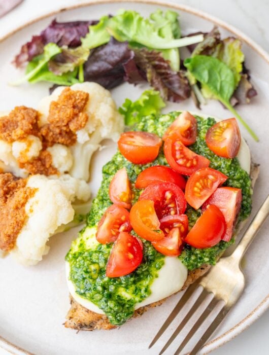 Chicken breast topped with mozarella, pesto, and tomatoes on a beige plate with cauliflower and a salad.
