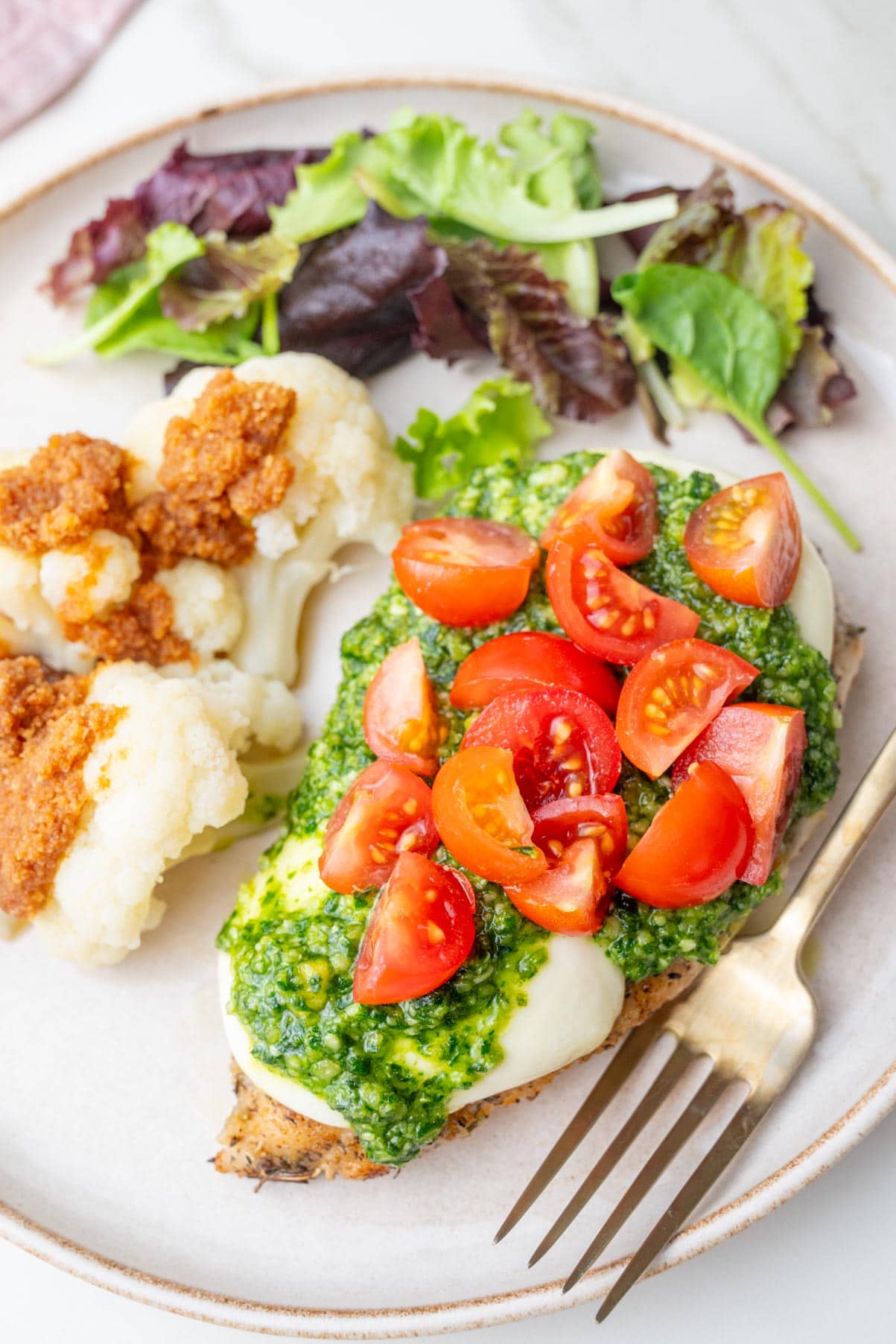 Chicken breast topped with mozarella, pesto, and tomatoes on a beige plate with cauliflower and a salad.