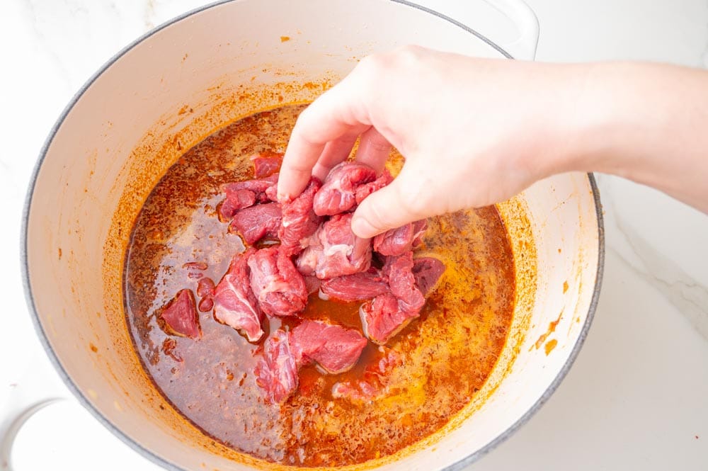 Raw beef cubes are being added to a pot with goulash.