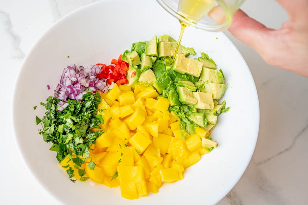 Olive oil is being added to a white bowl with chopped mango, avocado, red onion, and cilantro.