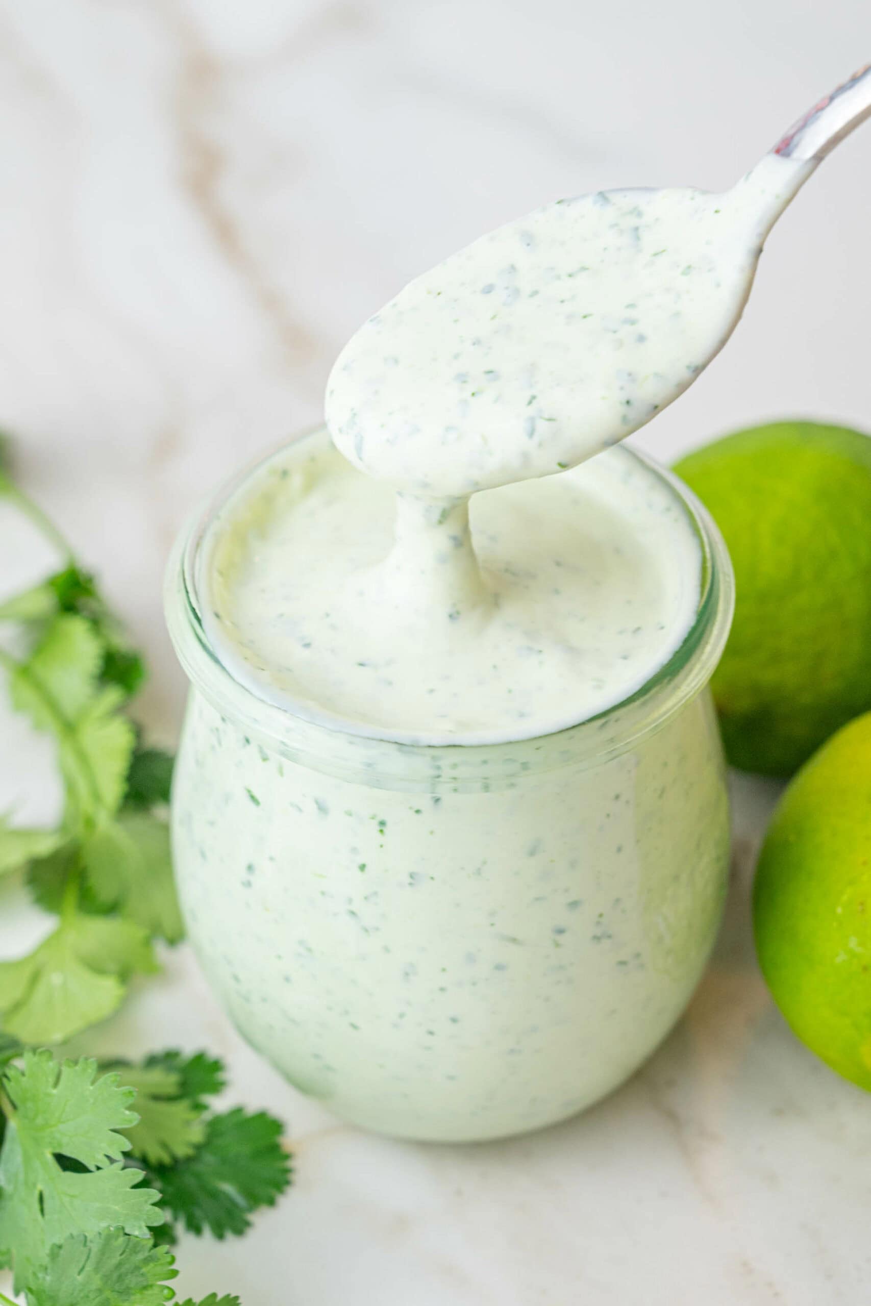 Cilantro Lime Crema is being drizzled from a spoon to a jar. Limes and cilantro in the background.