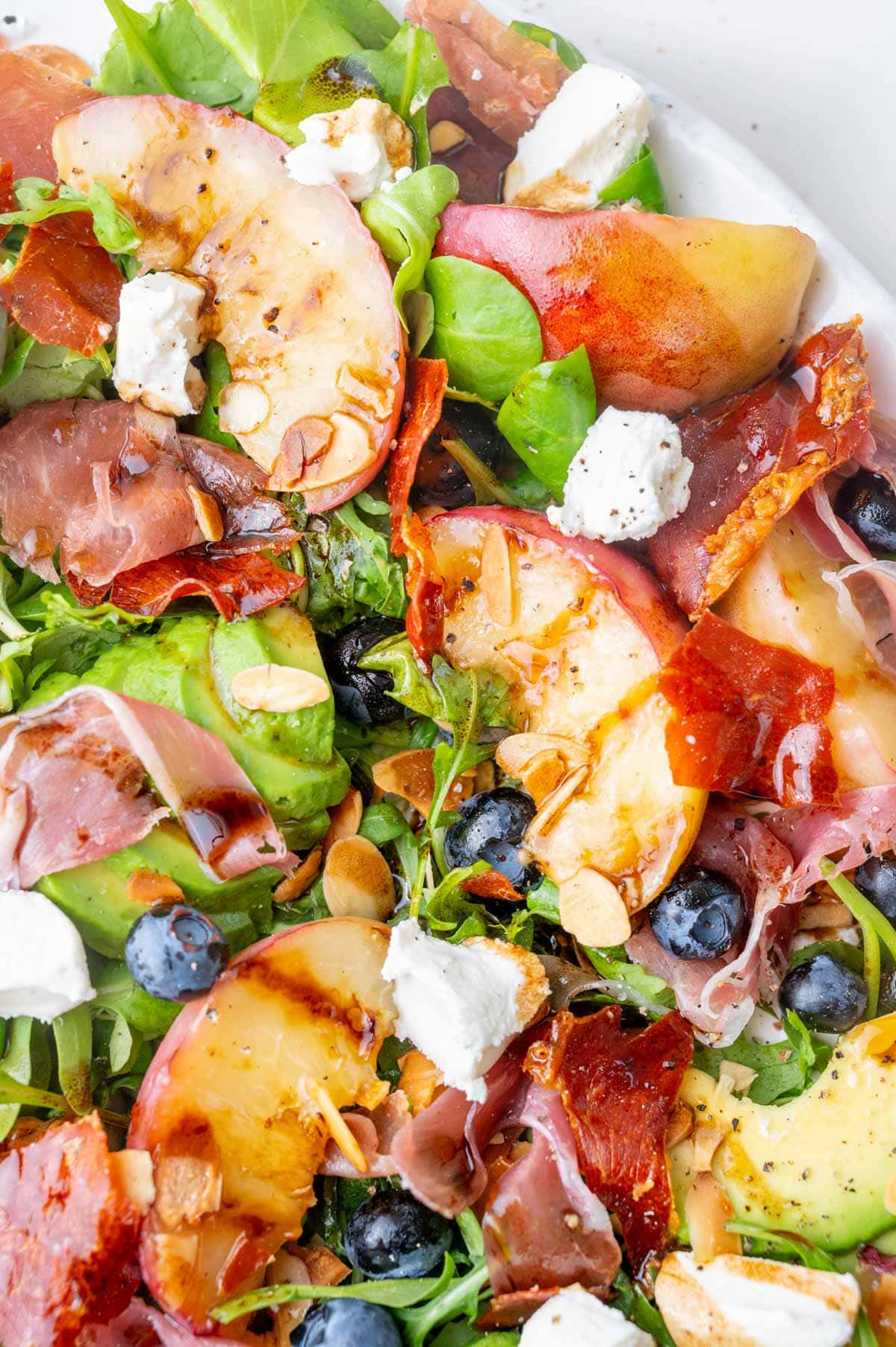 A close-up photo of Peach salad with prosciutto, blueberries, almonds, avocado, and goat cheese on a white plate.