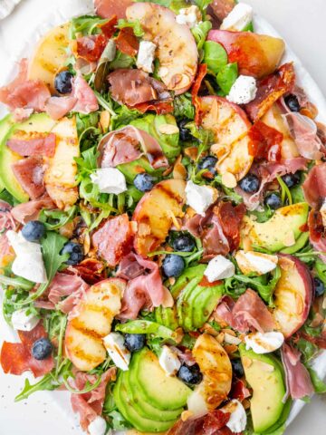 Peach salad with prosciutto, blueberries, almonds, avocado, and goat cheese on a white plate.