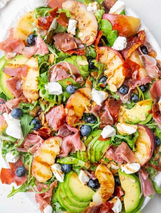Peach salad with prosciutto, blueberries, almonds, avocado, and goat cheese on a white plate.