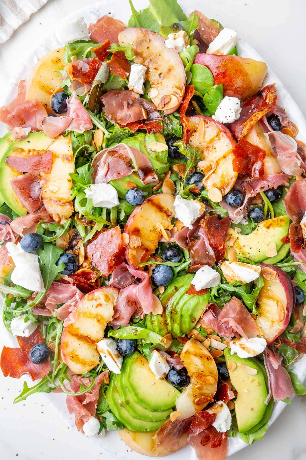 Peach salad with prosciutto, blueberries, almonds, avocado, and goat cheese on a white plate.