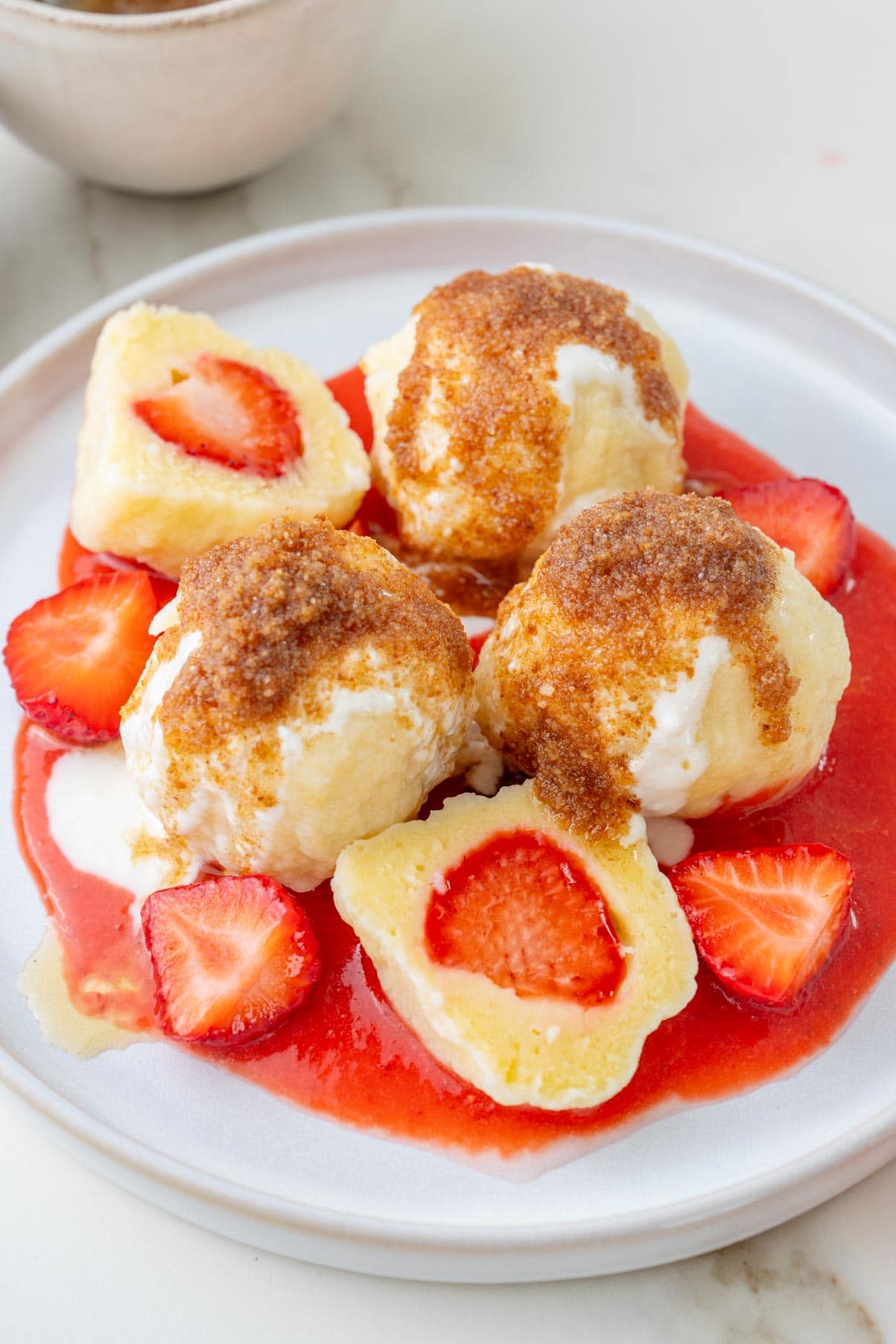 Polish strawberry dumplings served with fried breadcrumbs, sweetened sour cream, and strawberry sauce on a white plate.