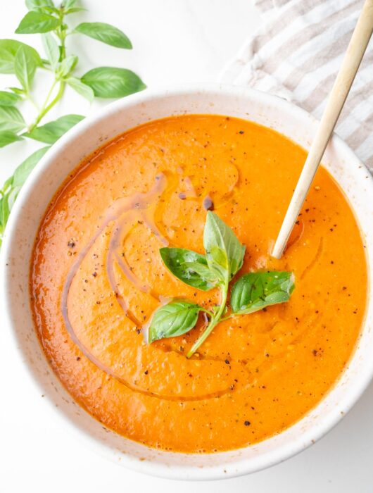 Roasted tomato soup in a white bowl topped with basil leaves and drizzled with olive oil.