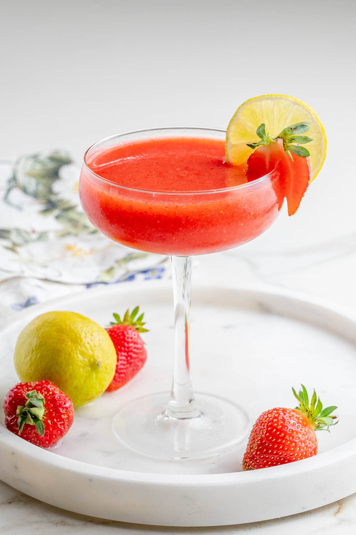 One glass with a strawberry margarita in a coupe glass garnished with a strawberry and a lime slice on a white stone board.