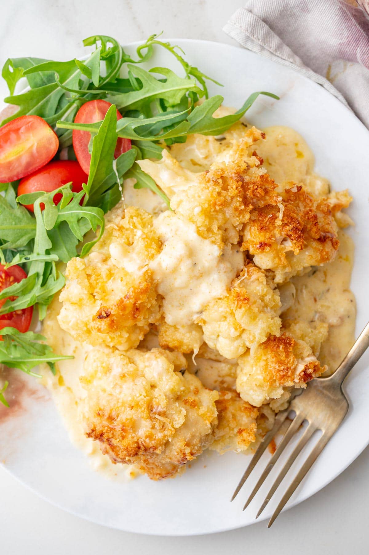 A serving of cauliflower gratin on a white plate with a side of arugula and tomatoes salad.