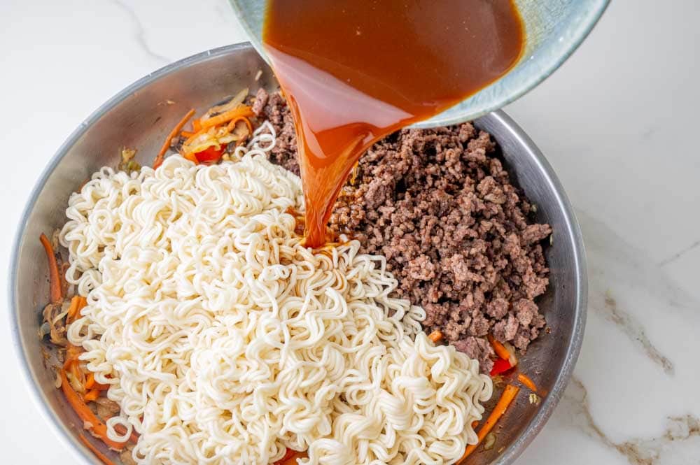 Sauce is being added to a pan with noodles, ground beef, and vegetables.