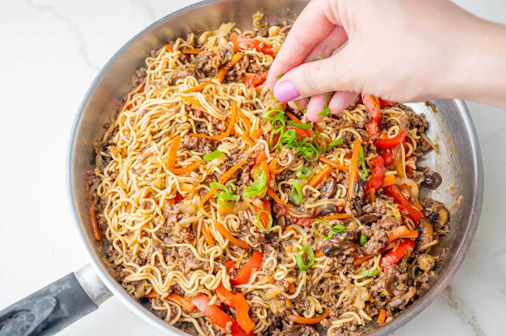 Sliced green onions are being sprinkled over ground beef noodles with vegetables.