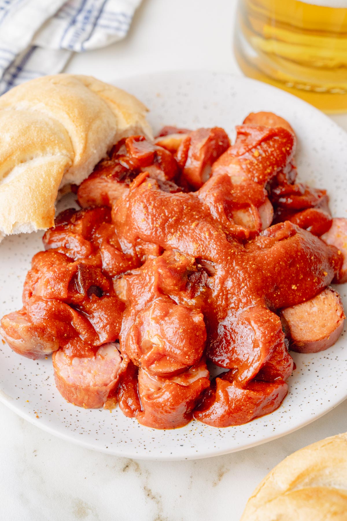 Currywurst on a white plate with bread rolls and beer in the background.