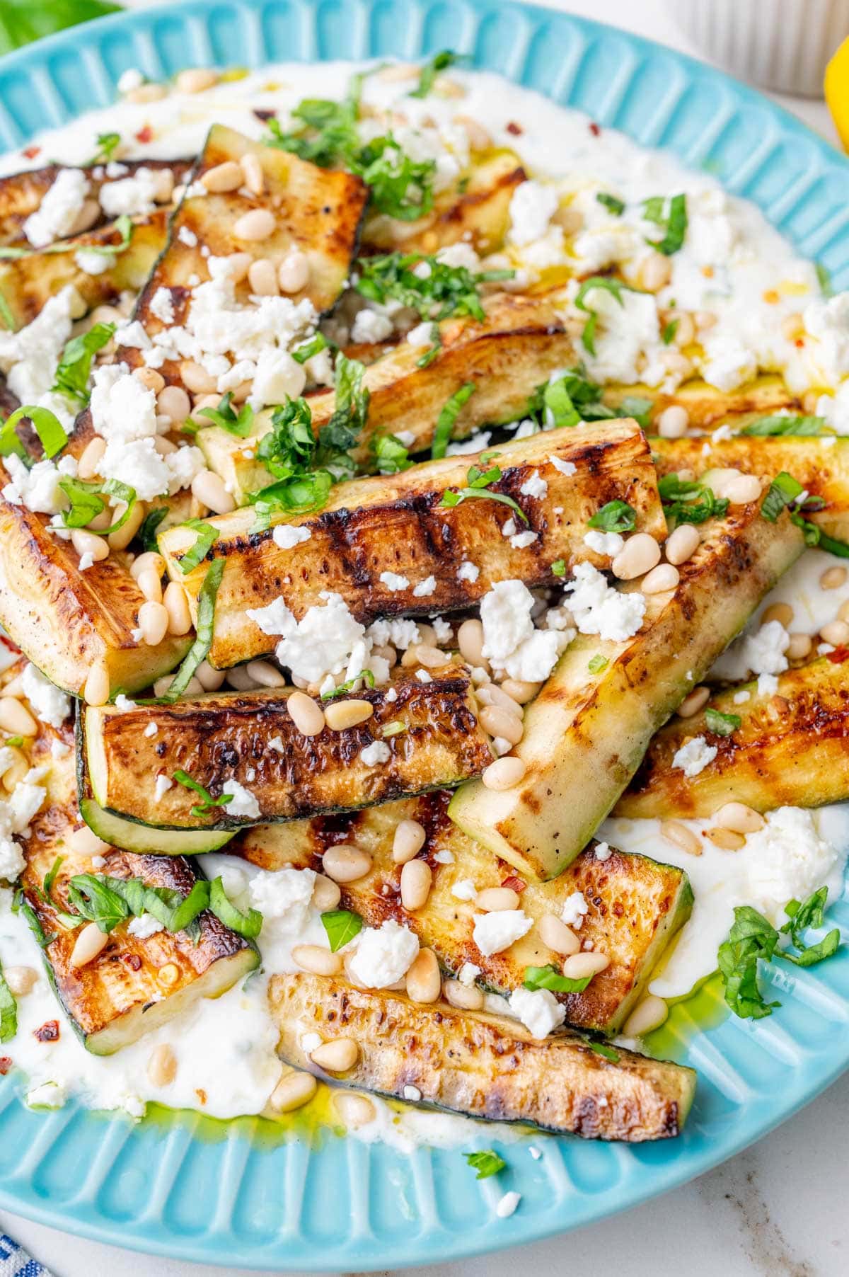 Grilled zucchini with yogurt, pine nuts, basil, and feta on a blue plate.