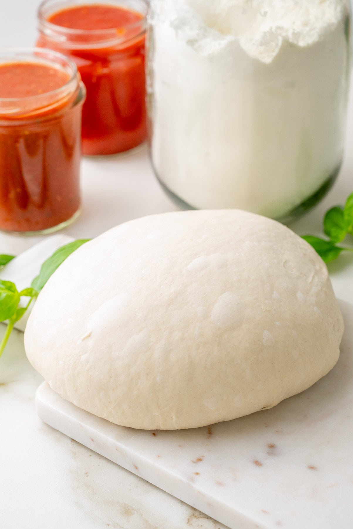 Pizza dough on a marbe board. Jars with tomato sauce and flour in the background.