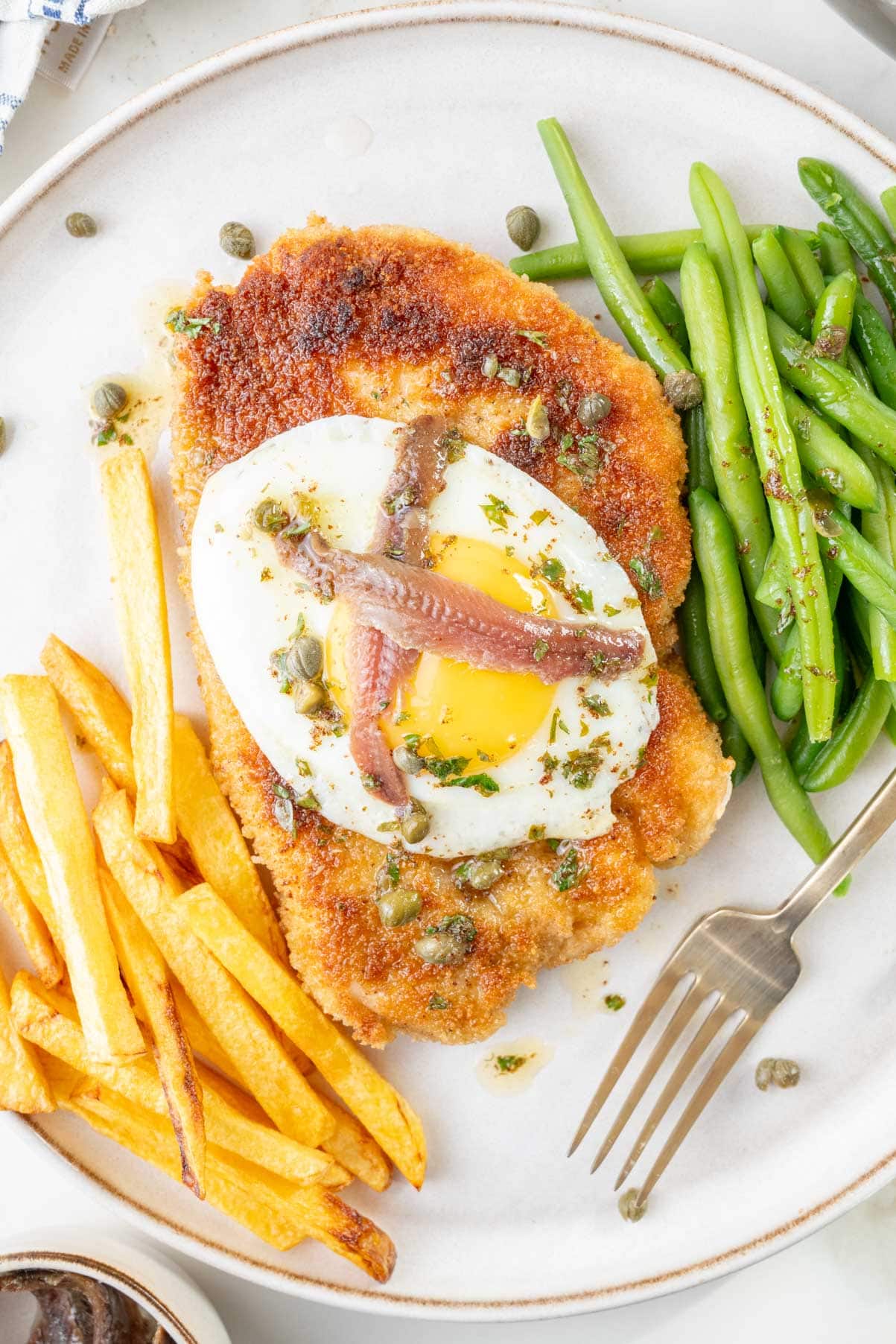 Schnitzel Holstein topped with fried egg and anchovies, served with fries and green beans on a white plate.