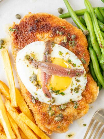 Schnitzel Holstein topped with fried egg, anchovies, and capers on a plate.