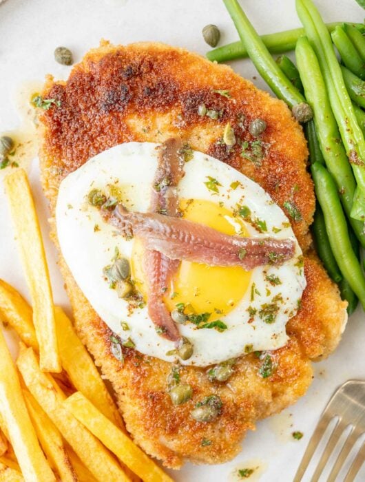 Schnitzel Holstein topped with fried egg, anchovies, and capers on a plate.