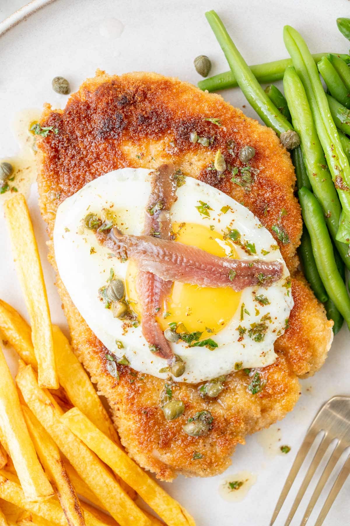 A close-up photo of Schnitzel Holstein topped with fried egg and anchovies and served with fries and green beans.