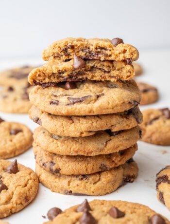 A stack of golden-brown Biscoff chocolate chip cookies, with the top cookie broken in half to show gooey chocolate inside. Other cookies are scattered in the background.