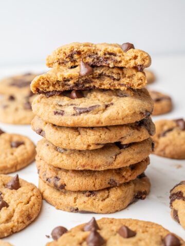 A stack of golden-brown Biscoff chocolate chip cookies, with the top cookie broken in half to show gooey chocolate inside. Other cookies are scattered in the background.