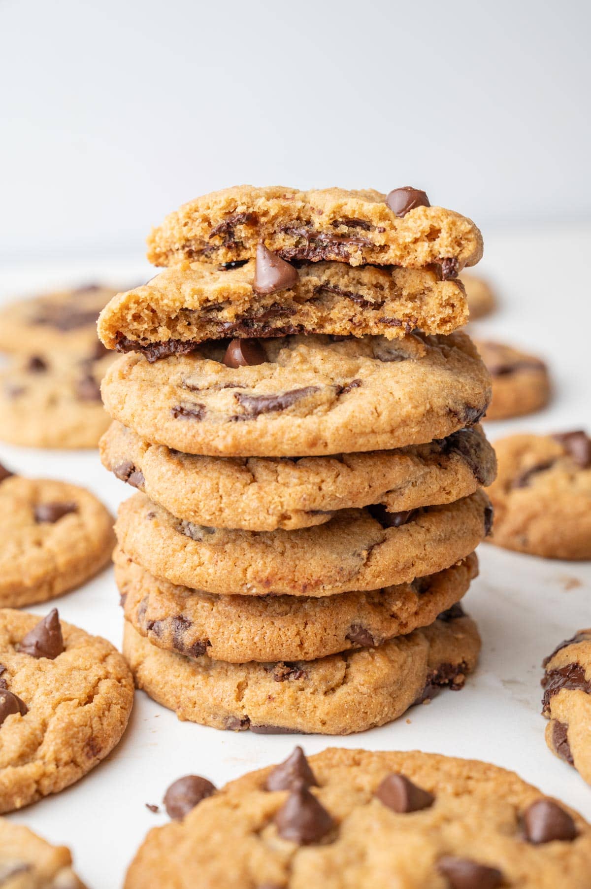 A stack of golden-brown Biscoff chocolate chip cookies, with the top cookie broken in half to show gooey chocolate inside. Other cookies are scattered in the background.