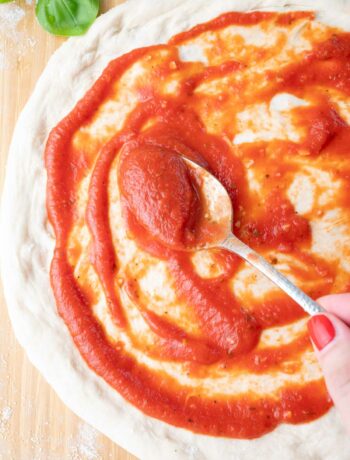 A hand uses a spoon to spread pizza sauce over a round pizza dough on a floured wooden surface, with fresh basil leaves in the top corner.