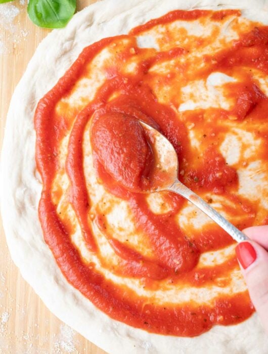 A hand uses a spoon to spread pizza sauce over a round pizza dough on a floured wooden surface, with fresh basil leaves in the top corner.