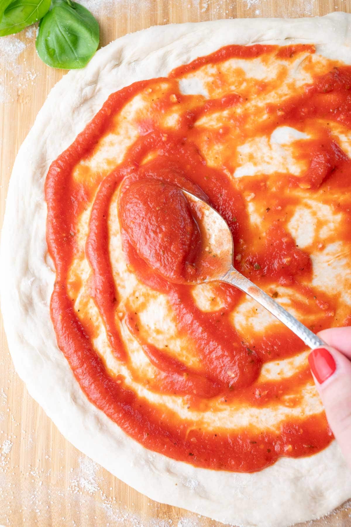 Pizza sauce is being spread on a pizza dough on a wooden board.