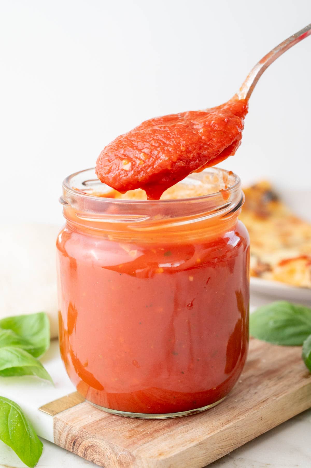 A spoon lifts a thick, bright red pizza sauce from a glass jar, with fresh basil leaves and a partially visible pizza in the background.