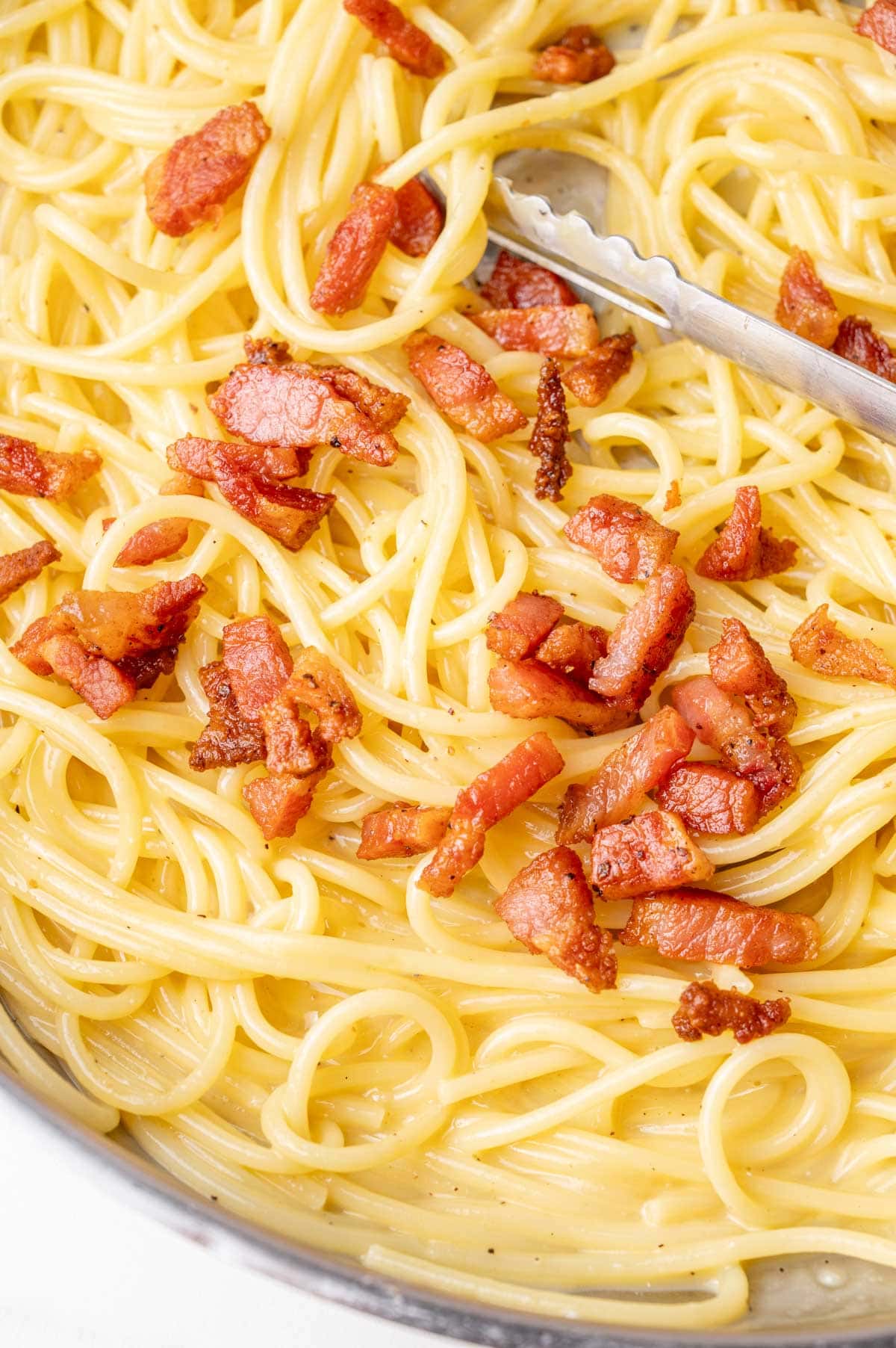 A close-up of a pan filled with spaghetti carbonara, featuring golden pasta coated in a creamy sauce and topped with crispy, browned guanciale pieces. A pair of metal tongs rests on the side.