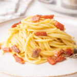 A close-up of a plate of spaghetti carbonara, featuring golden pasta coated in a creamy sauce with crispy chunks of guanciale and a sprinkle of black pepper. A fork rests at the side, with a pan of additional carbonara visible in the blurred background.