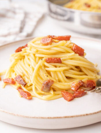 A close-up of a plate of spaghetti carbonara, featuring golden pasta coated in a creamy sauce with crispy chunks of guanciale and a sprinkle of black pepper. A fork rests at the side, with a pan of additional carbonara visible in the blurred background.