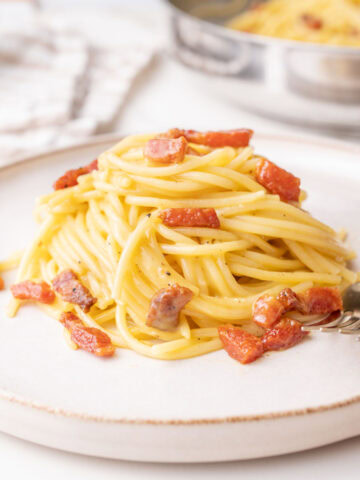 A close-up of a plate of spaghetti carbonara, featuring golden pasta coated in a creamy sauce with crispy chunks of guanciale and a sprinkle of black pepper. A fork rests at the side, with a pan of additional carbonara visible in the blurred background.