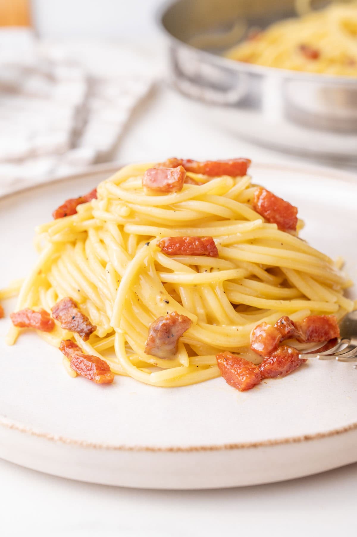 A close-up of a plate of spaghetti carbonara, featuring golden pasta coated in a creamy sauce with crispy chunks of guanciale and a sprinkle of black pepper. A fork rests at the side, with a pan of additional carbonara visible in the blurred background.