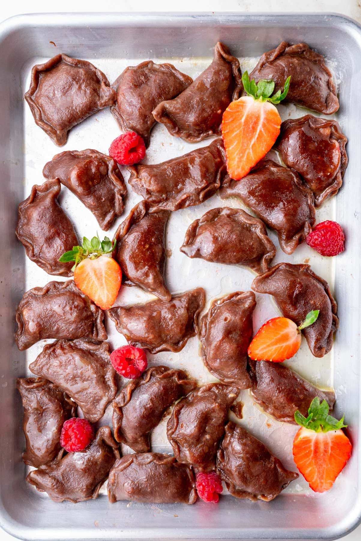 A tray of freshly cooked chocolate pierogi, made with cocoa-flavored dough and arranged on a metal baking sheet. The dumplings have a glossy surface and are garnished with fresh strawberries and raspberries.