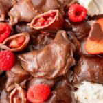 A close-up photo of chocolate pierogi served on a plate, showing their strawberry and raspberry filling. The dumplings are drizzled with chocolate sauce and accompanied by fresh berries and a dollop of whipped cream.