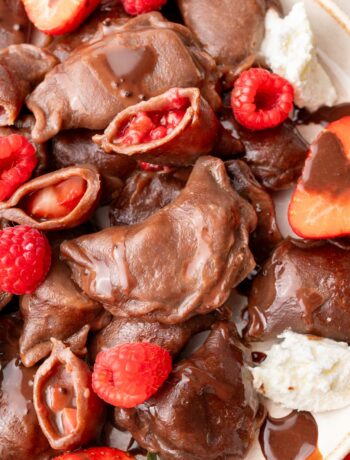 A close-up photo of chocolate pierogi served on a plate, showing their strawberry and raspberry filling. The dumplings are drizzled with chocolate sauce and accompanied by fresh berries and a dollop of whipped cream.