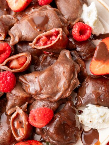 A close-up photo of chocolate pierogi served on a plate, showing their strawberry and raspberry filling. The dumplings are drizzled with chocolate sauce and accompanied by fresh berries and a dollop of whipped cream.