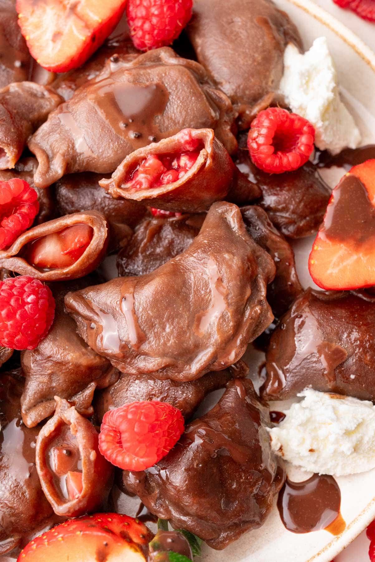 A close-up of chocolate pierogi served on a plate, showing their strawberry and raspberry filling. The dumplings are drizzled with chocolate sauce and accompanied by fresh berries and whipped cream.