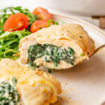 A close-up of a spinach-stuffed chicken breast, sliced in half to reveal a creamy spinach and cheese filling. The chicken is golden brown, covered with melted cheese, and served with a side of fresh arugula and cherry tomatoes.