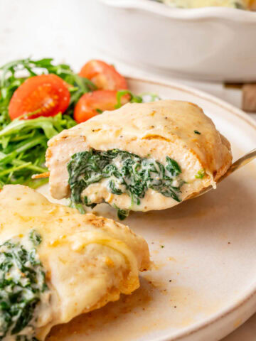 A close-up of a spinach-stuffed chicken breast, sliced in half to reveal a creamy spinach and cheese filling. The chicken is golden brown, covered with melted cheese, and served with a side of fresh arugula and cherry tomatoes.