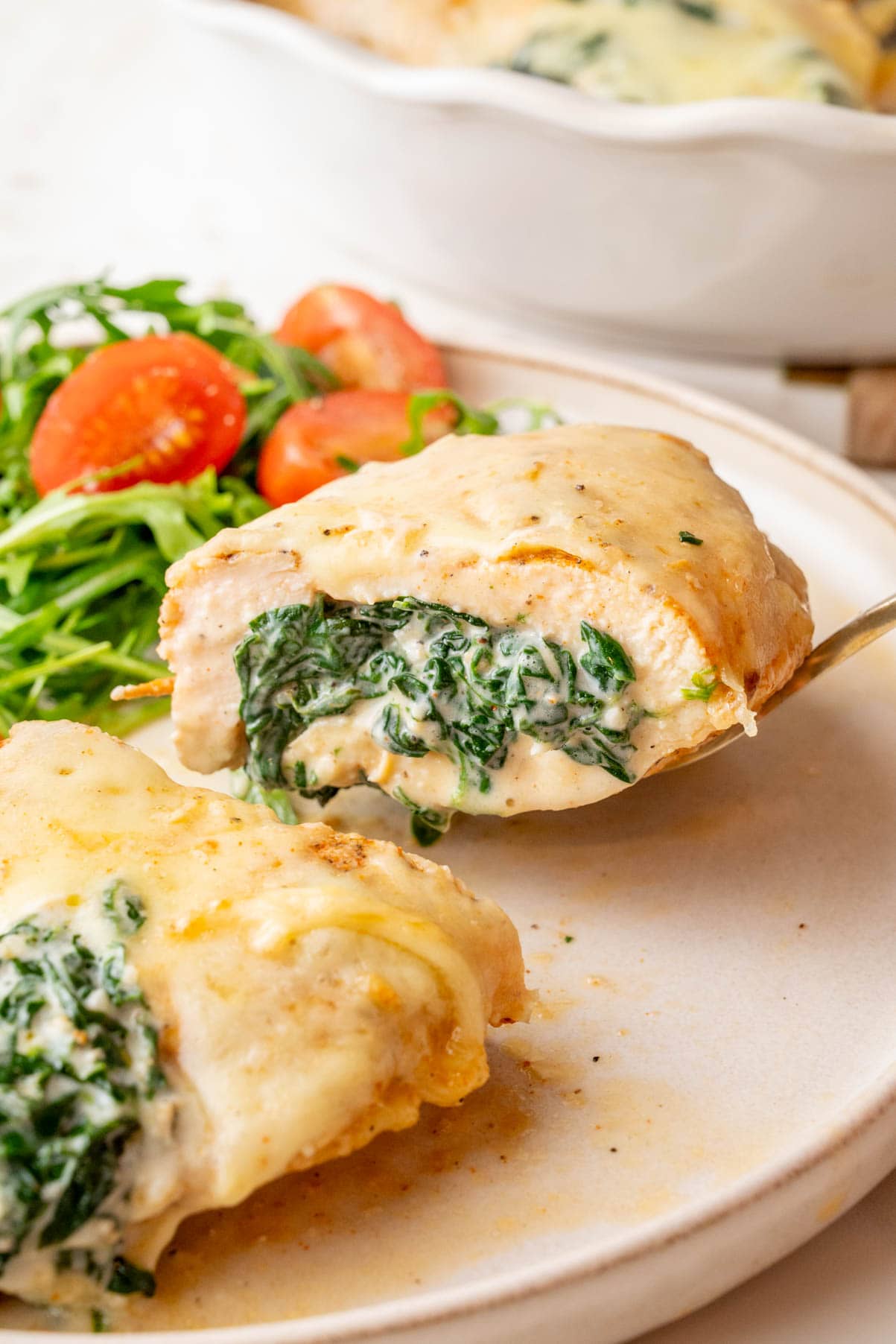 A close-up of a spinach-stuffed chicken breast, sliced in half to reveal a creamy spinach and cheese filling. The chicken is golden brown, covered with melted cheese, and served with a side of fresh arugula and cherry tomatoes. 